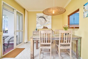 A dining area with a wooden table and chairs, a large painting on the wall, and a window with blue shutters.