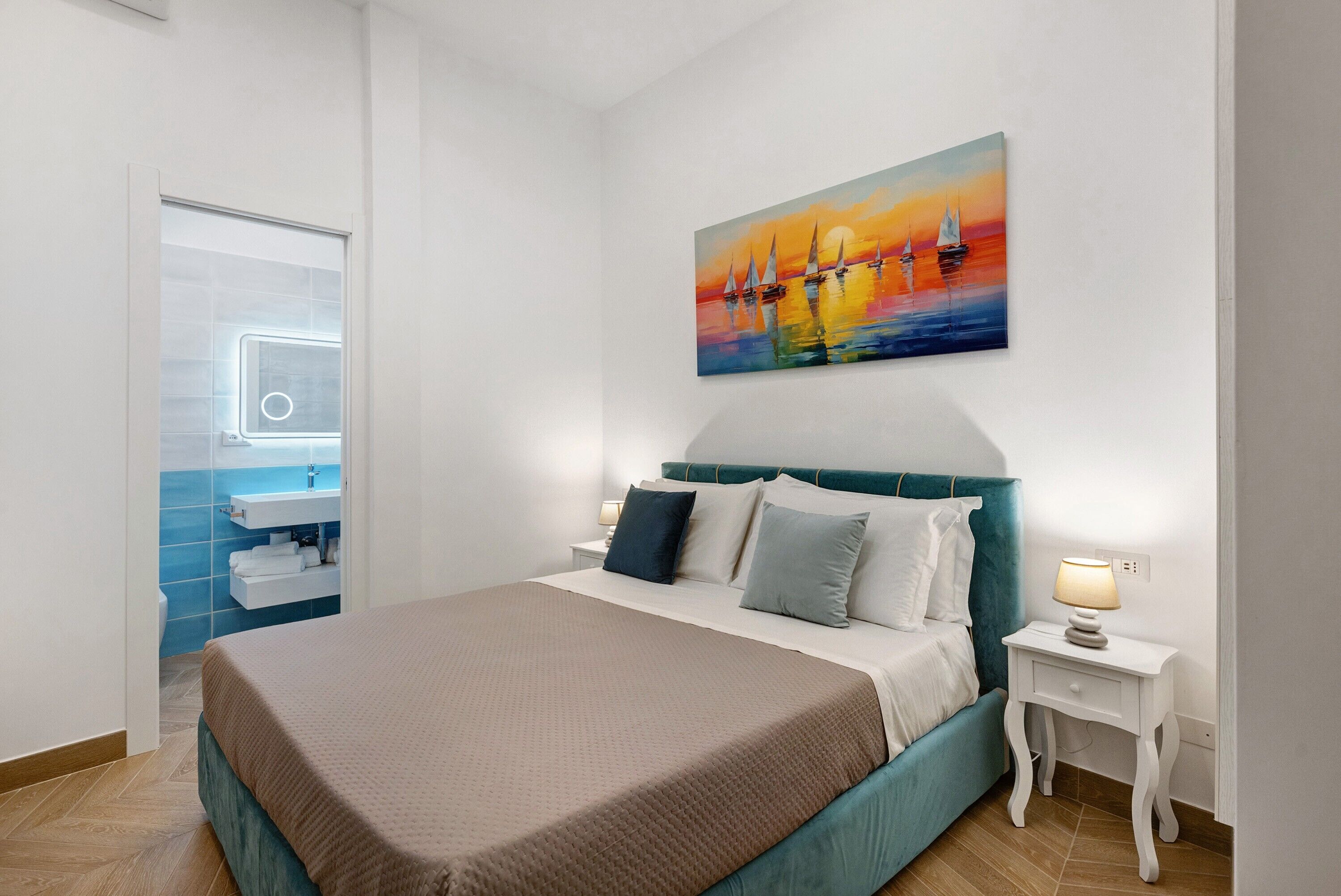 Ein modernes Schlafzimmer mit einem großen Bett, einem Segelboot-Gemälde, einem Badezimmer mit blauen Fliesen und einem weißen Nachttisch mit Lampe.