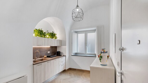 Una cocina moderna con armarios blancos, piso con diseño de espiga, ventana con cortinas e iluminación colgante.