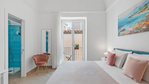 Un dormitorio con cama, una silla, un balcón y un cuadro.
