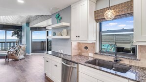 A modern kitchen with white cabinets, a granite countertop, and a large window overlooking the ocean. - Oceanfront Penthouse | 3BR/2BA | Pools, Beach Access & Incredible Views (Port Aransas)
