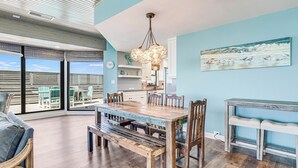 A spacious dining area with a wooden table and benches, a chandelier, and a view through large windows. - Oceanfront Penthouse | 3BR/2BA | Pools, Beach Access & Incredible Views (Port Aransas)