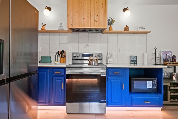 A modern kitchen with blue cabinets, stainless steel appliances, and wooden shelves.