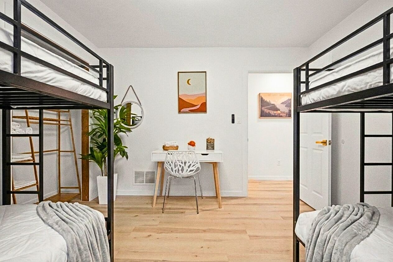 A dormitory room with bunk beds, a desk, a chair, a potted plant, and artwork on the walls.