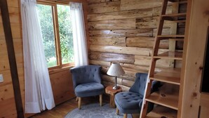 Une pièce chaleureuse avec des murs en bois, un tapis rond, deux fauteuils bleus, une petite table d’appoint en bois avec une lampe, et une étagère en bois en forme d’échelle.