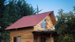 Une cabane en bois avec un toit rouge, une véranda avec deux chaises et un petit arbre à feuilles persistantes dans le jardin.