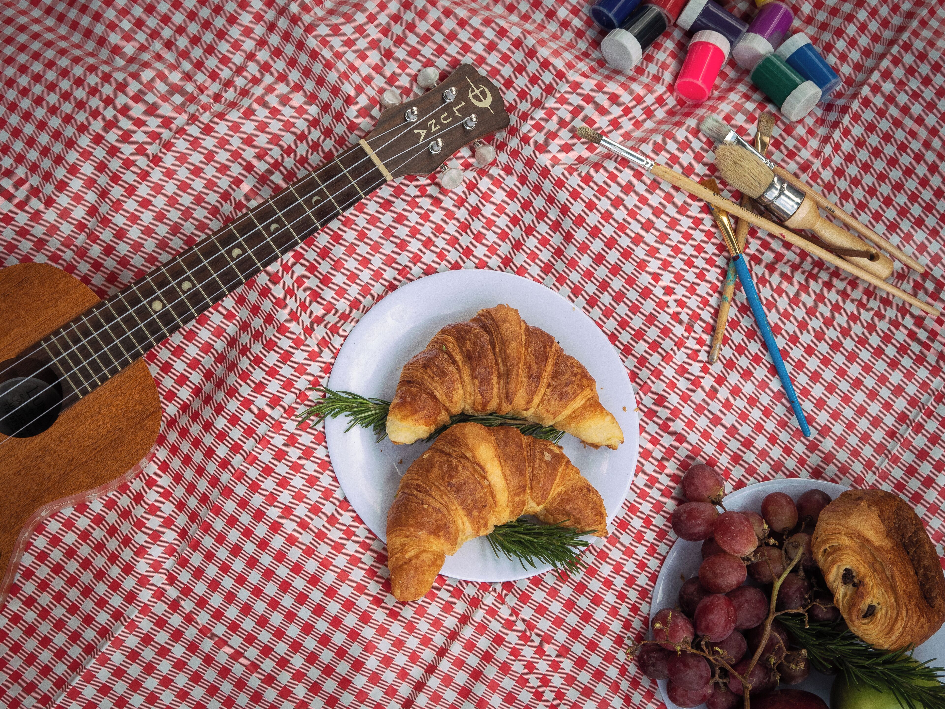 Una guitarra, croissants, raïms i pinzells sobre un mantell daurat a quadres.
