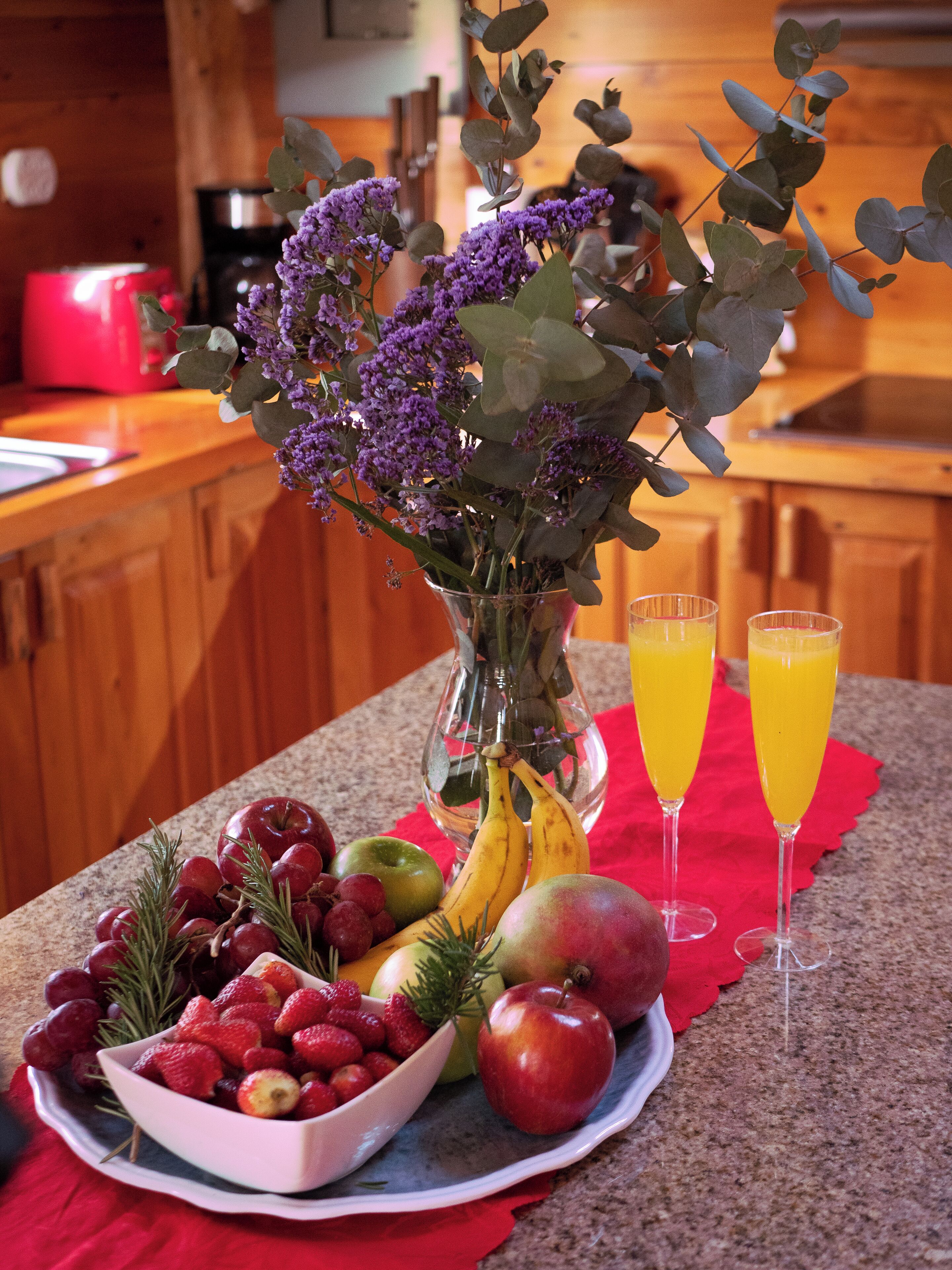 A fruit bowl with grapes, strawberries, and apples, a vase with purple flowers, and two glasses of orange juice on a kitchen counter.