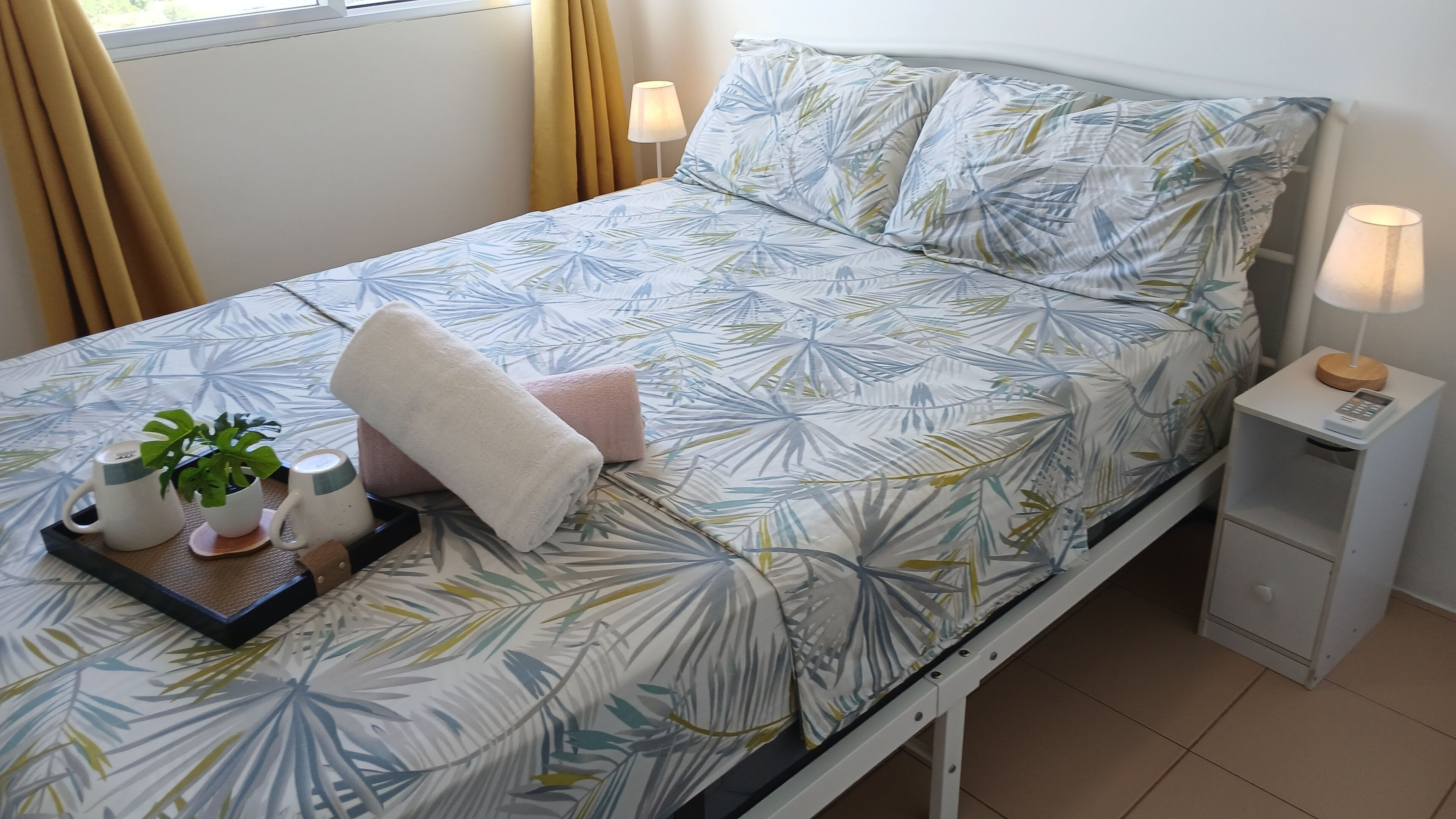 A neatly made bed with a tropical leaf pattern bedspread, two pillows, a bedside lamp, a small bedside table with a lamp, and a tray with a plant and a mug.