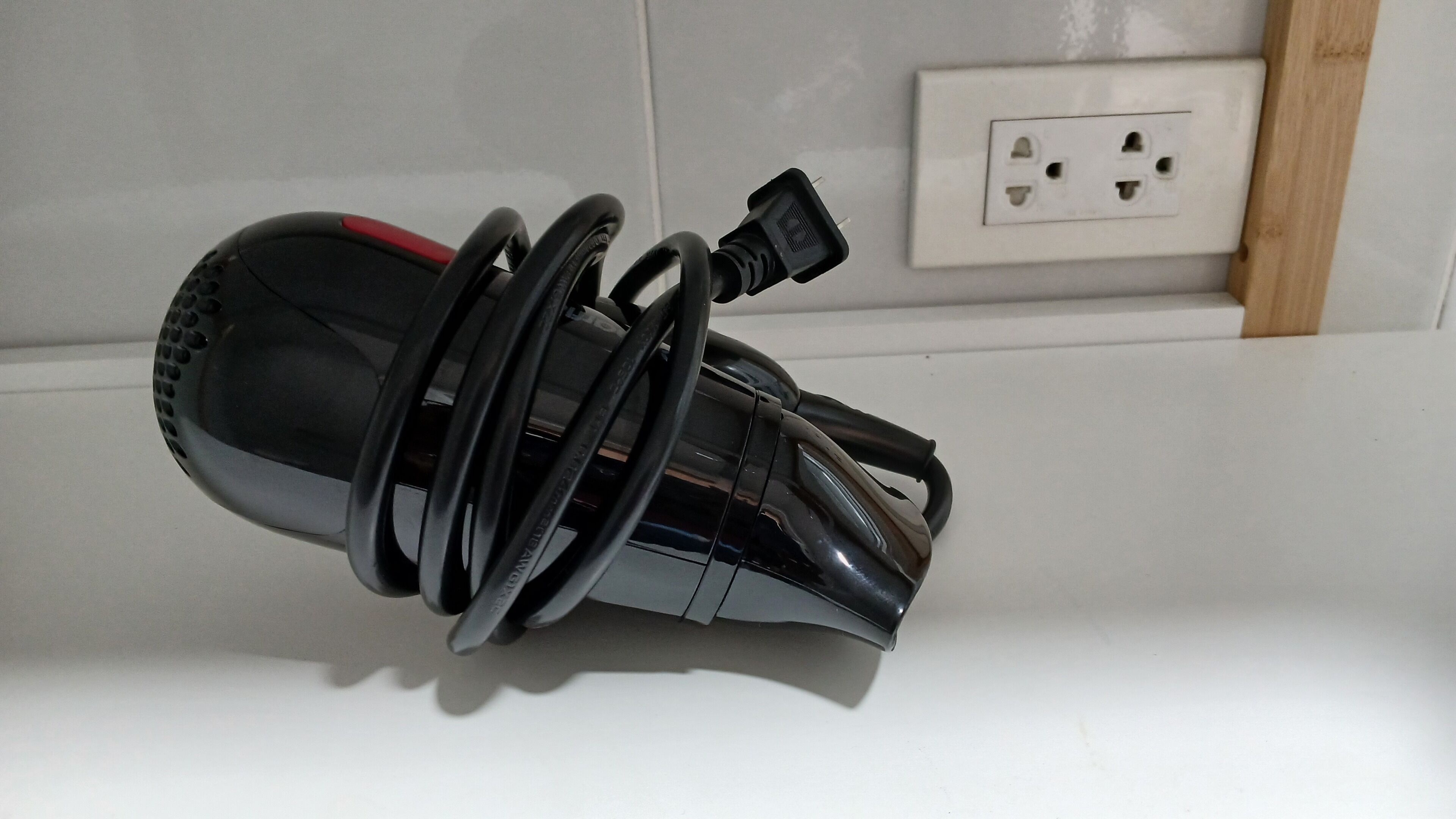A black, cylindrical object with a red stripe, coiled and resting against a white wall near a power outlet.