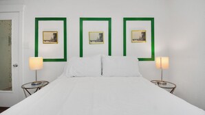 A hotel room with a large bed, white linens, green-framed pictures on the wall, and two bedside lamps.