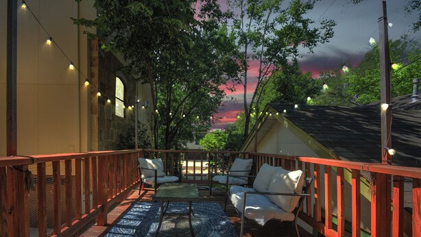A covered outdoor seating area with a patterned rug, wooden railings, and string lights.