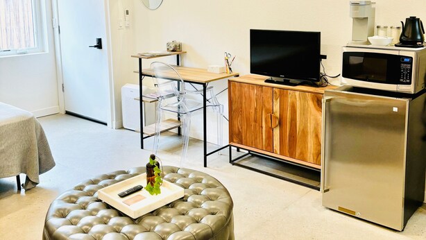 A modern, well-lit room with a flat-screen TV, a coffee maker, a microwave, a refrigerator, and a seating area with a tufted ottoman.