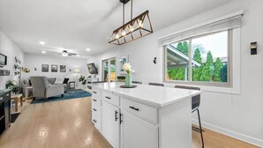 A modern kitchen with white cabinetry, a central island, and a large window offering a view of greenery. - In-Ground Pool AND Hot Tub! Fenced Yard, Pet Friendly Home. Near Town! (Crystal Lake)