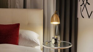 Ein ordentlich bezogenes Bett mit roter und weißer Bettwäsche, ein kleiner runder Tisch mit einer Lampe sowie eine Wand mit abstrakter Kunst.