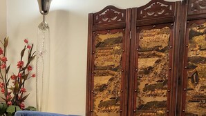 Ein blaues Sofa mit roten Kissen, ein Glastisch mit einem roten Topf und eine dekorative Holzvertäfelung mit goldenen Verzierungen.