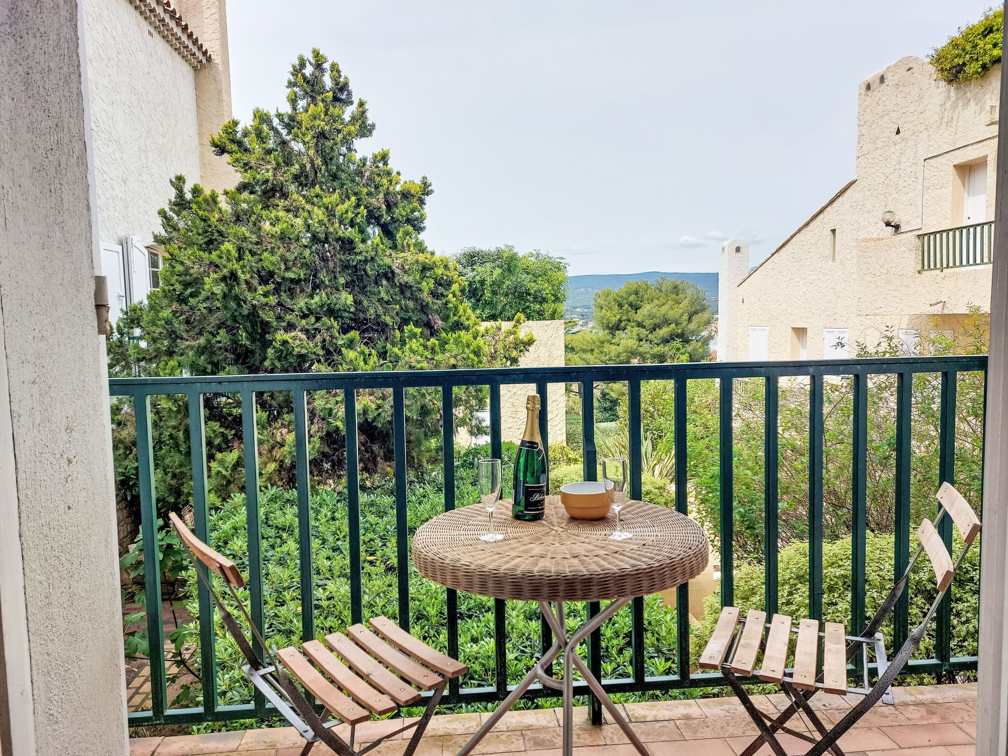 Un balcon avec une petite table ronde en osier et deux chaises en bois, une bouteille de champagne et un verre.