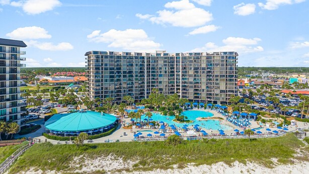Un hotel con varios pisos, un amplio espacio de piscina con sombrillas y una playa al frente.