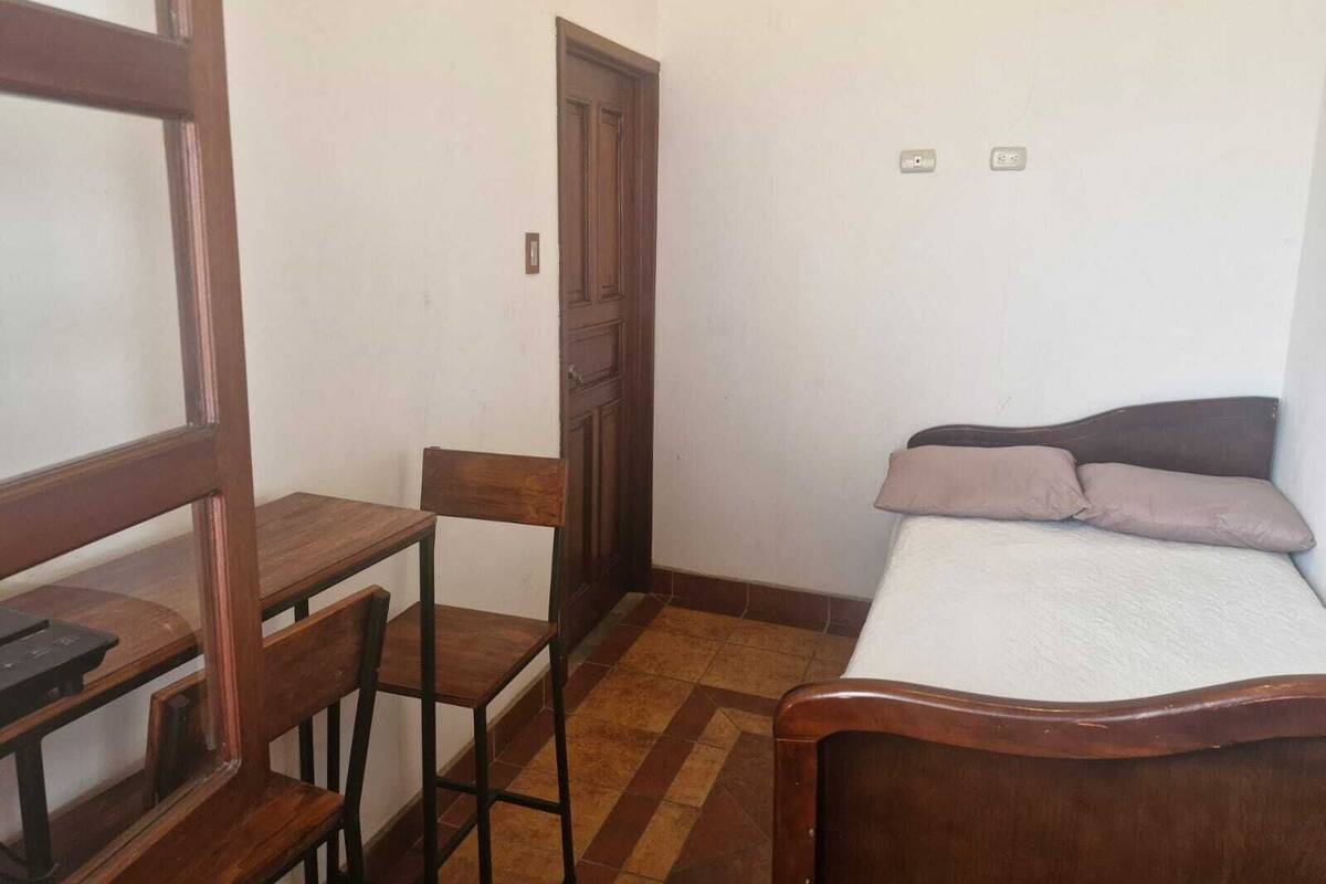A small, simple room with a wooden desk, two chairs, a bed with a headboard, and a door.