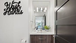 Ein modernes Badezimmer mit einem dunklen Holz-Waschtisch, einem weißen Waschbecken und einem Spiegel darüber. An der Wand befindet sich ein Schild mit der Aufschrift 'Wash Your Hands' und auf dem Waschtisch ein Blumentopf.