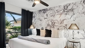 Ein Schlafzimmer mit einem großen Bett, dunklen Vorhängen, einem Deckenventilator, floralem Wandpapier und einem kleinen Tisch mit einer Lampe.