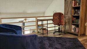 A room with a sloped ceiling, a blue armchair, a purple rug, a bookshelf, and a ceiling fan. - Charming 2-bedroom home with fenced yard sleeps 6 in Baton Rouge near Tigerland (Baton Rouge)