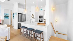 A modern kitchen with a central island, bar stools, stainless steel appliances, pendant lighting, and a view of the living area.