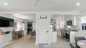 A modern kitchen with white cabinetry, a built-in microwave, and a large stove. There is a dining area with a table and chairs, and a television mounted on the wall. - Beach, Breeze & Bunk Beds. FamilyFun Steps to the Shore. Dog-Friendly, GameWall. (Wareham)