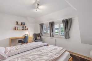 Une chambre à coucher avec un lit, un bureau, une chaise, un radiateur et une fenêtre avec des rideaux.