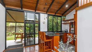 A modern kitchen with a wooden ceiling, large windows, a built-in oven, and a dining area with wooden chairs. - NATURAVE HOME - Tiny house with mountain view (Monteverde)