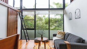A modern living room with a grey sofa, wooden floor, a round coffee table, and a view of greenery through large windows. - NATURAVE HOME - Tiny house with mountain view (Monteverde)