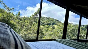 A bed with a view of a lush, green hillside through large windows. - NATURAVE HOME - Tiny house with mountain view (Monteverde)