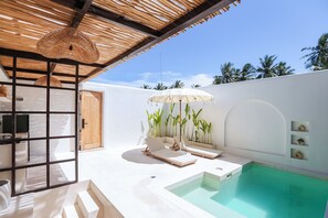 Un área de piscina moderna al aire libre con dos sillas de descanso, una piscina pequeña y un techo de paja.