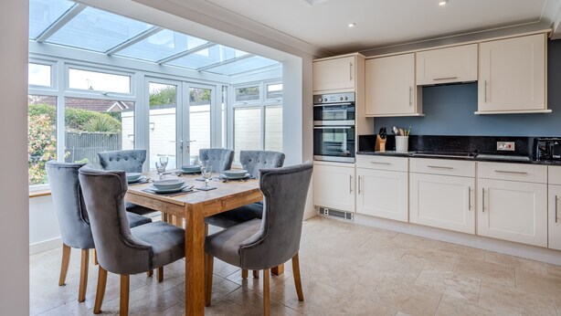 A modern kitchen with a dining area, featuring a large window, built-in oven, and white cabinetry. - Bosuns Cottage | Dog Friendly Coastal Cottage, 5 Minutes from the Beach (Winterton-on-sea)