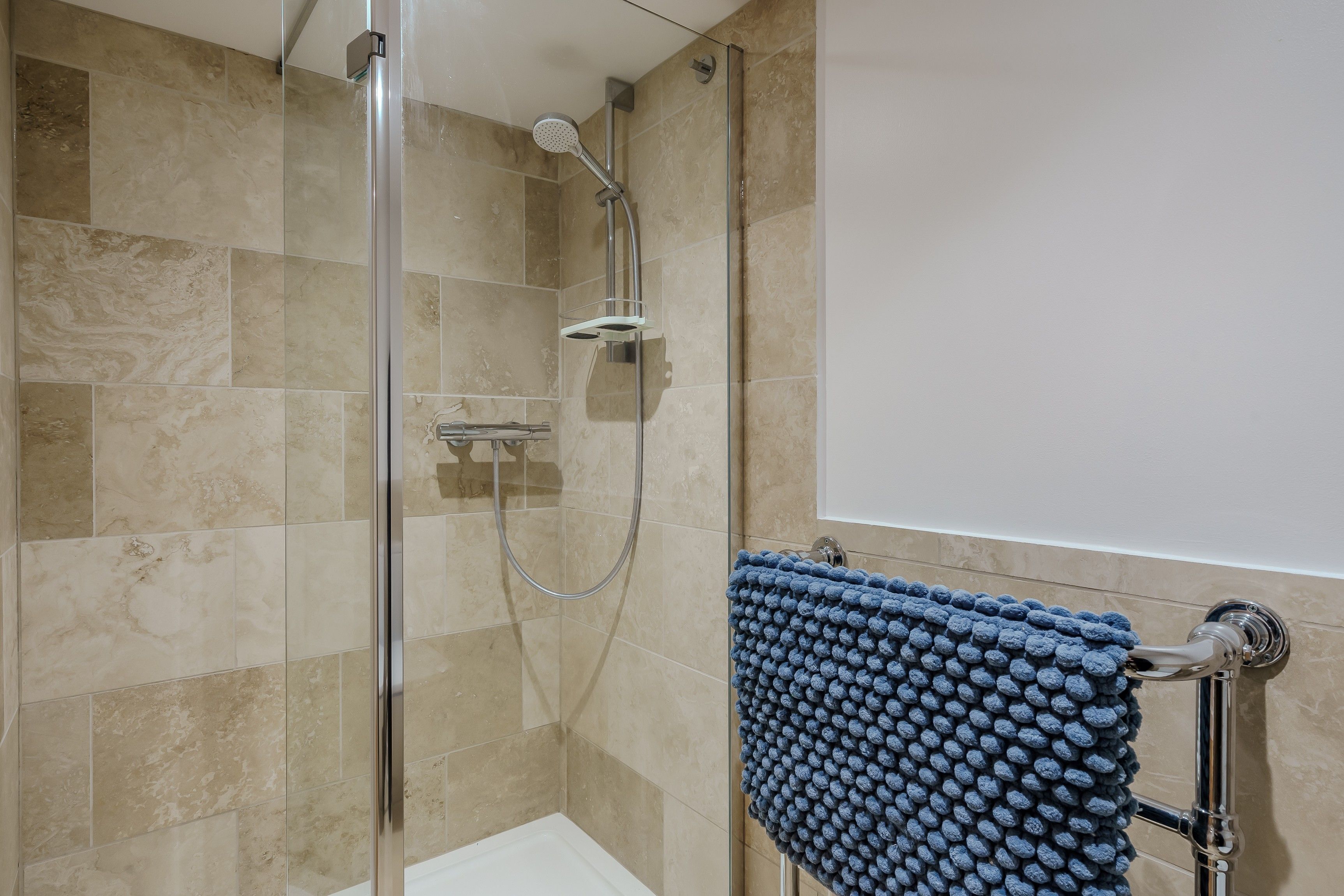 Ducha con puerta de vidrio, una toalla azul con textura y azulejos color beige.