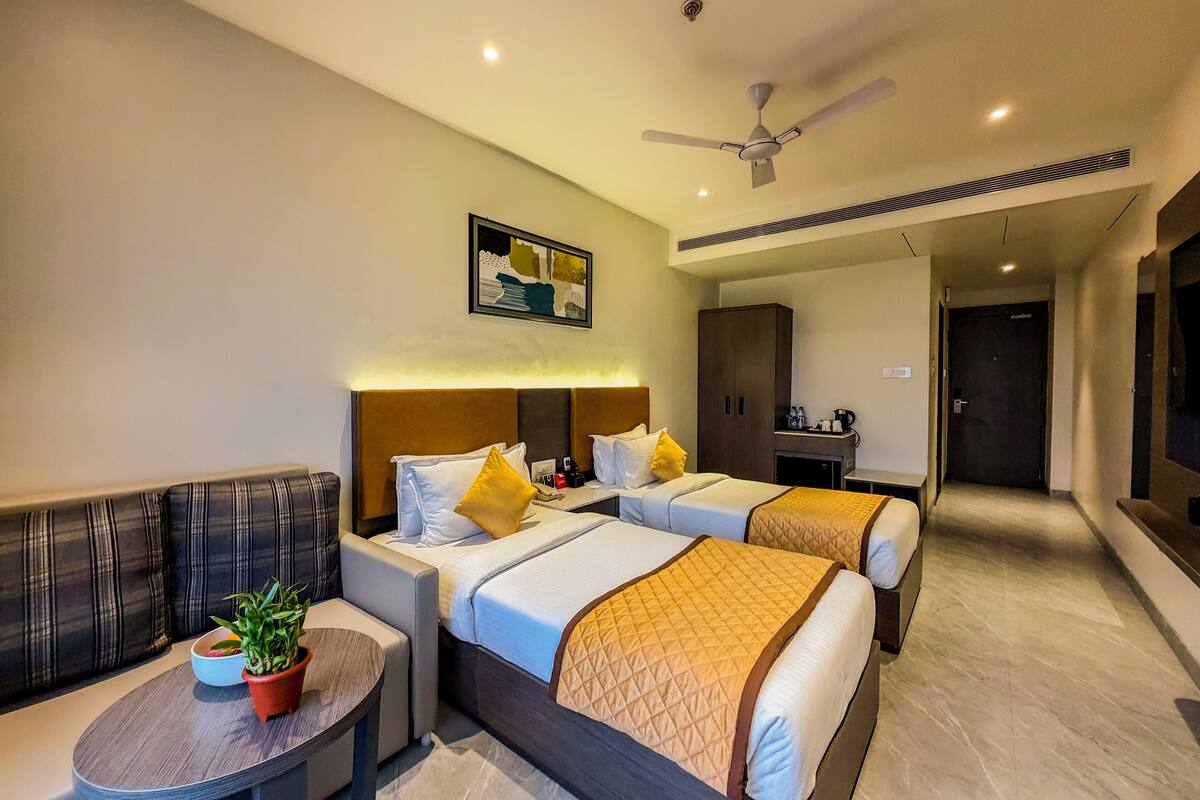 A hotel room with two beds, a sofa, a small table with a plant, a ceiling fan, and a framed picture on the wall.