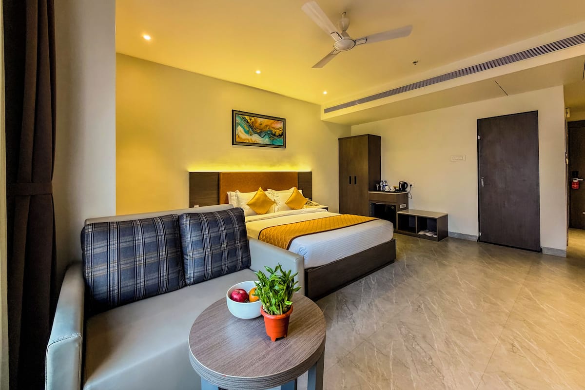 A hotel room with a bed, a sofa, a small table with a plant, a ceiling fan, and a painting on the wall.