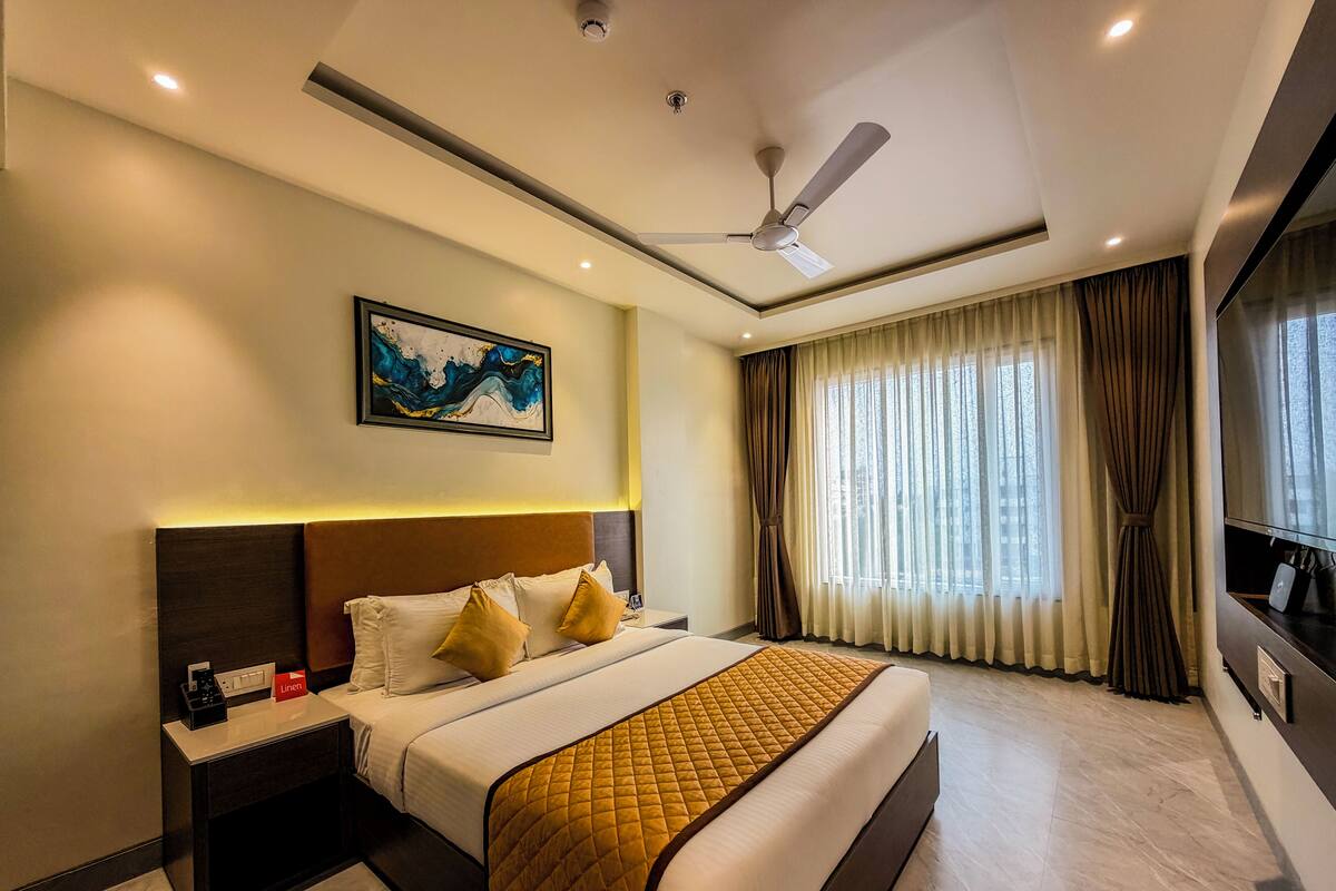 A hotel room with a large bed, a ceiling fan, a painting, a TV, and a window with curtains.