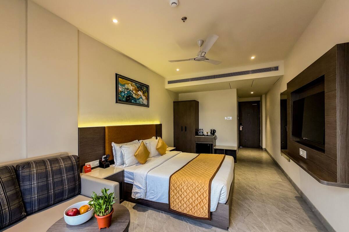 A hotel room with a bed, a sofa, a small table with a bowl of fruit and a plant, a ceiling fan, and a TV mounted on the wall.