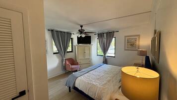 Une chambre à coucher comprenant un lit, une chaise, un ventilateur de plafond, une fenêtre avec des rideaux et une lampe.