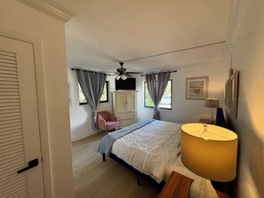 Et soveværelse med seng, stol, loftventilator, et vindue med gardiner og en lampe.
