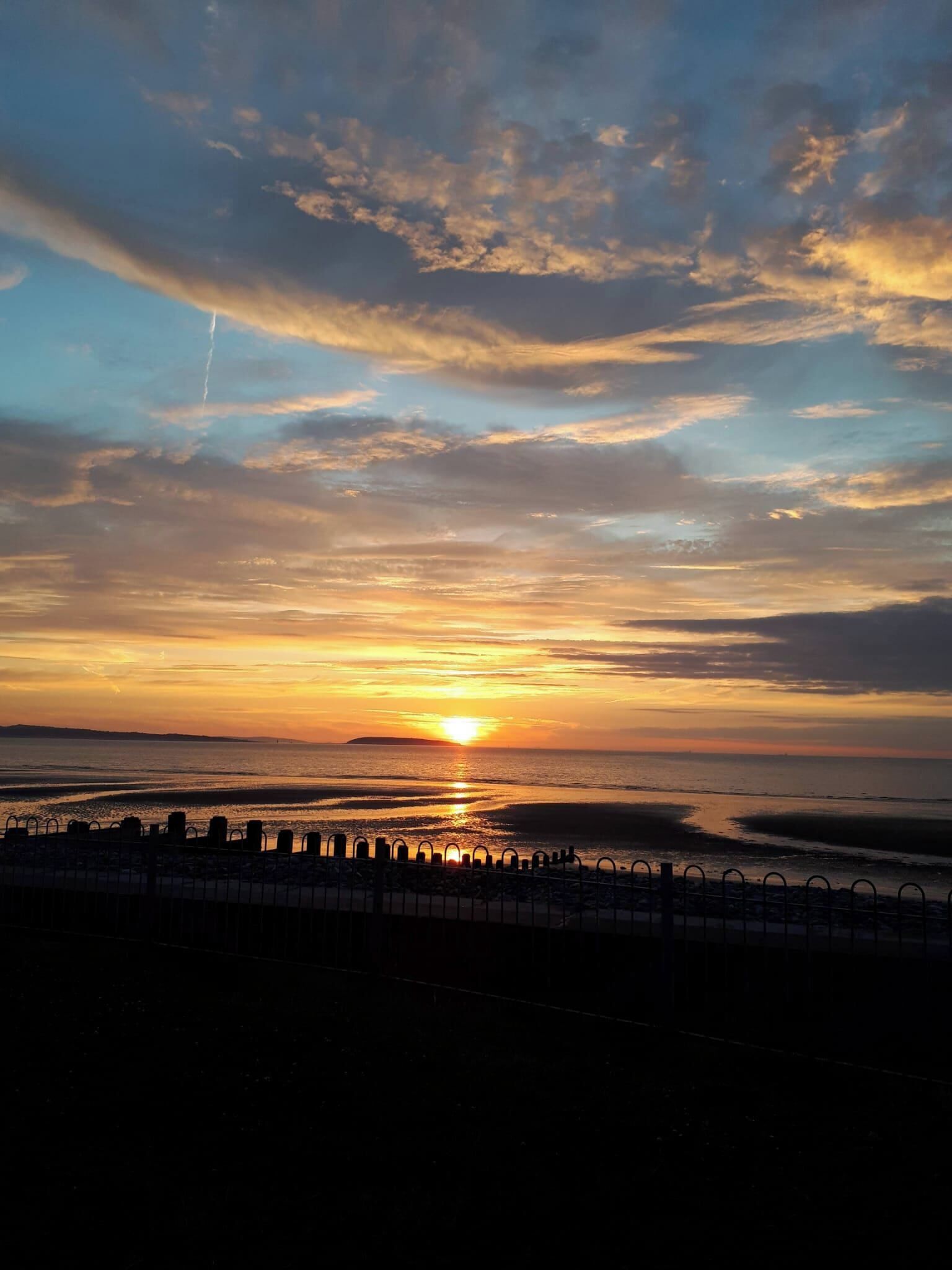 Een zonsondergang boven een wateroppervlak, met een metalen hek in de voorgrond.