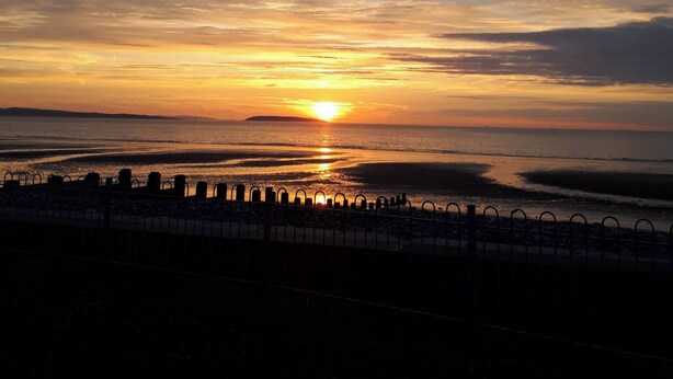 A sunset over a body of water with a metal fence in the foreground.