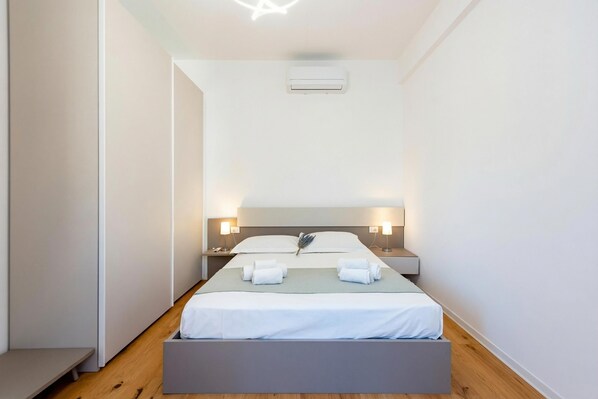 A modern hotel room with a bed, bedside tables, a bench, and an air conditioning unit.