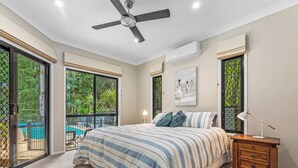 A bedroom with a bed, a ceiling fan, a nightstand, and a window with a view of greenery. - Toogoom Beach Vibes. Absolute Beachfront Home with pool! 4BR (Toogoom)