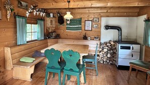 A rustic cabin interior with a wooden dining table, two green chairs, a bench, a stove, and various decorations including antlers and framed pictures. - Christoph by Interhome (Haus)