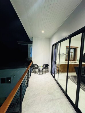 Un balcón con baranda de madera, una pequeña mesa con sillas y vistas al interior a través de grandes puertas de cristal.