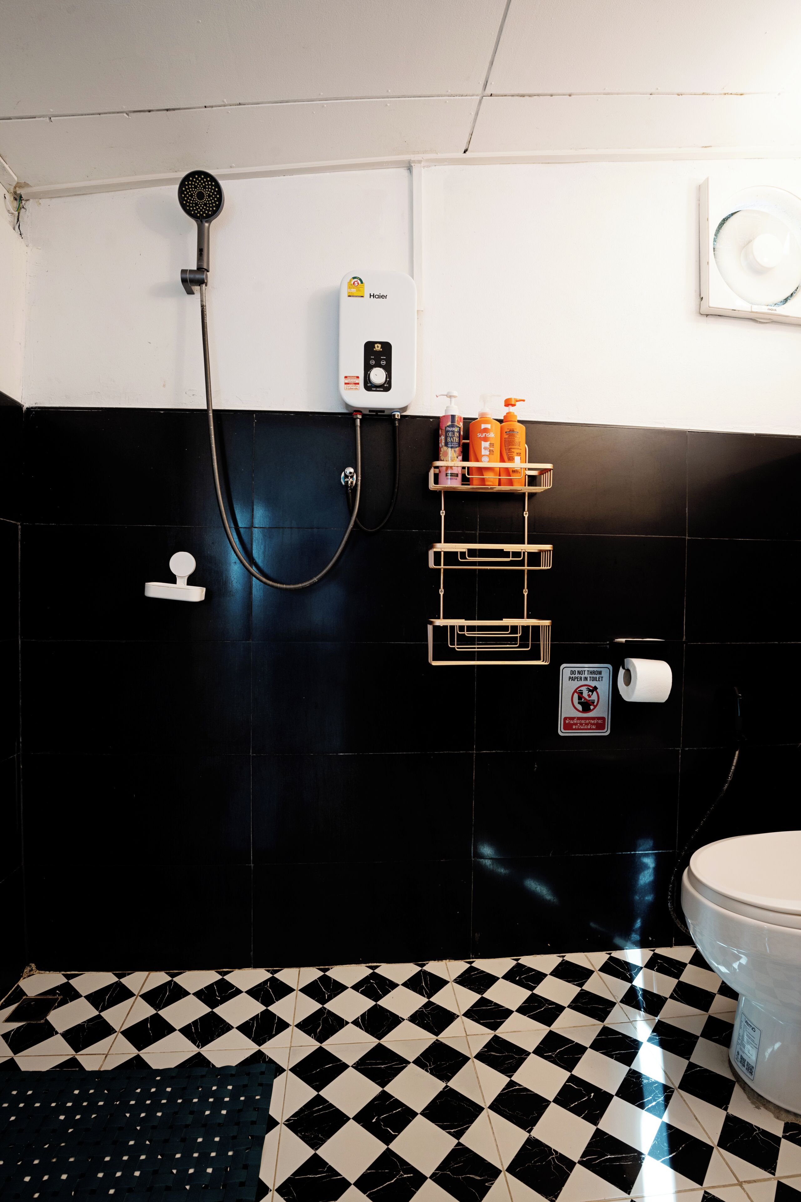 A bathroom with black and white checkered floor tiles, a shower with a handheld showerhead, a soap dispenser, and a toilet.