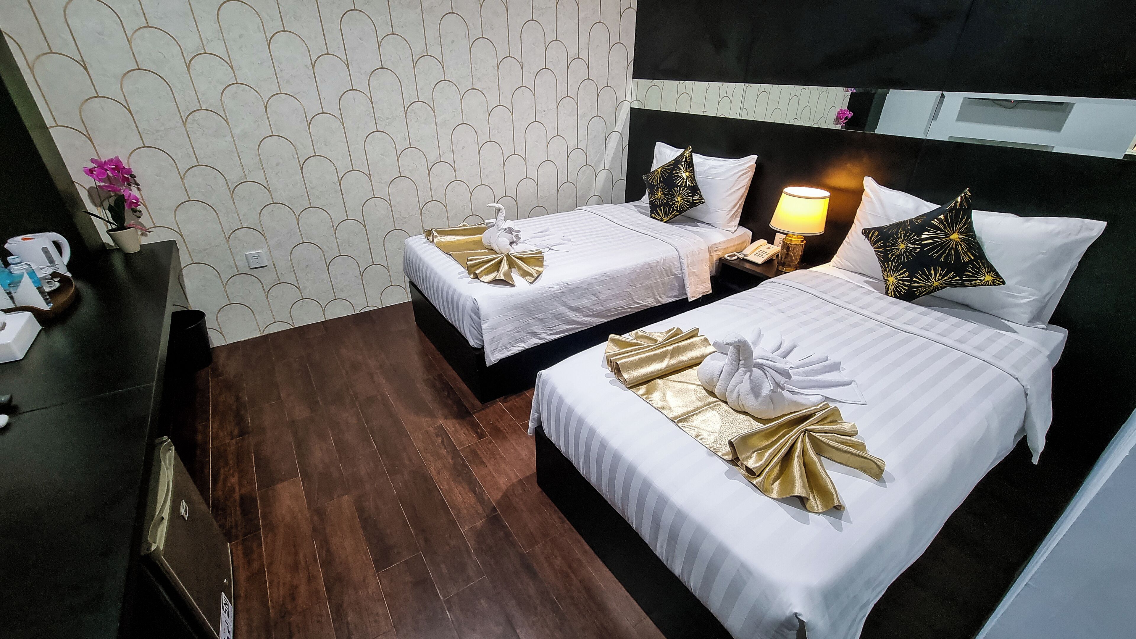 A hotel room with two beds, each with white bedding and gold accents, a bedside table with a lamp, and a patterned wall.