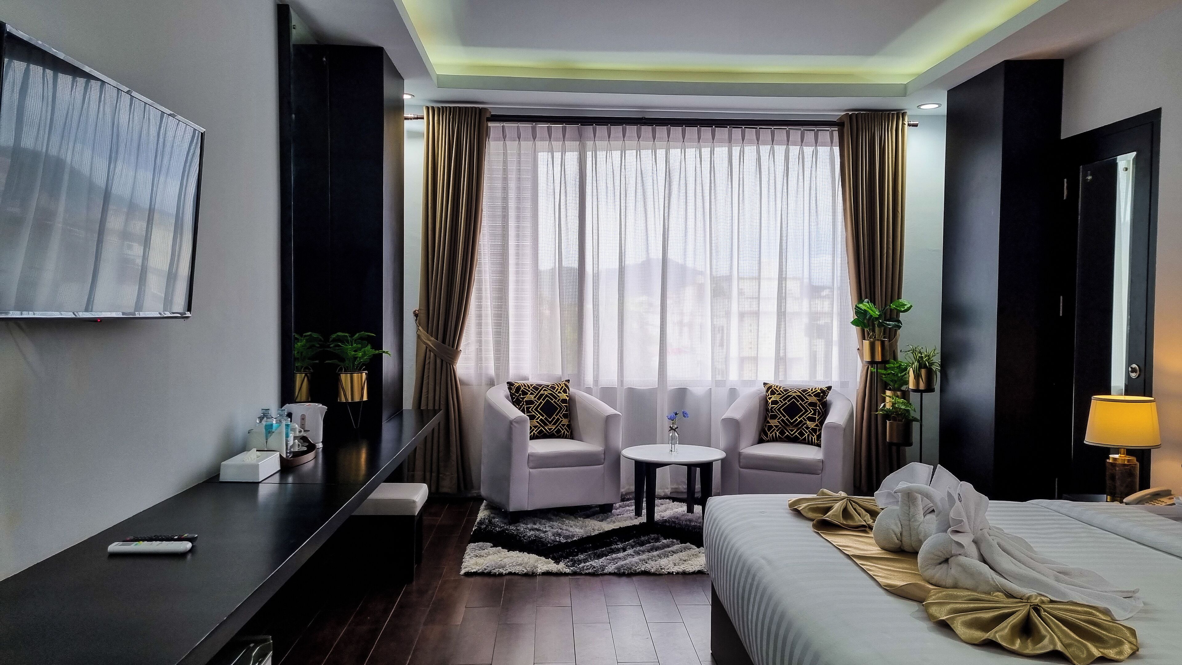 Une chambre d’hôtel moderne, équipée d’un lit, de deux fauteuils, d’une petite table, d’un téléviseur et d’une grande fenêtre avec des rideaux.